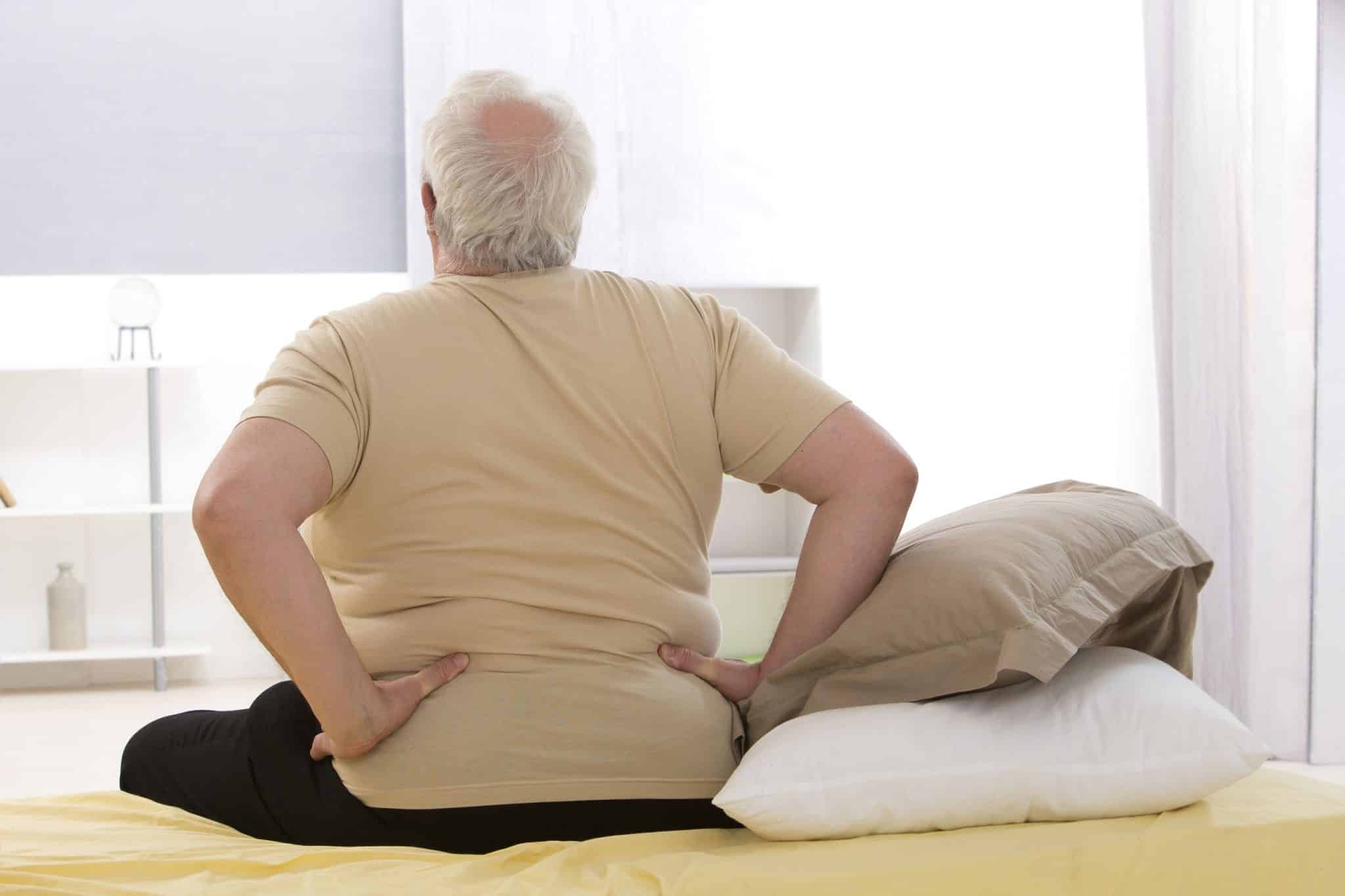Senior man holding lower back while sitting on the edge of a bed