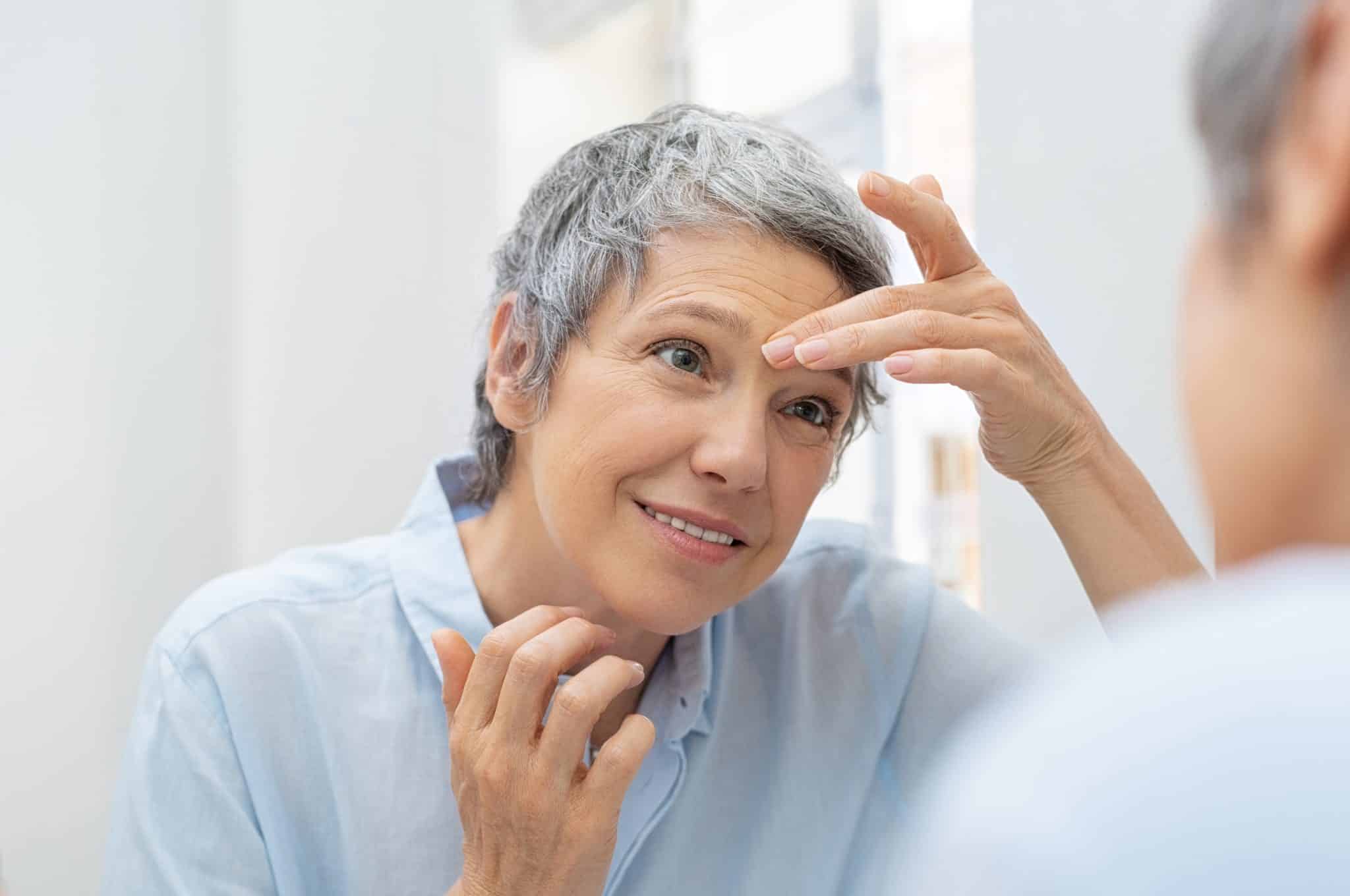 Older woman smiling and looking her face in the bathroom mirror while touching wrinkles on forehead