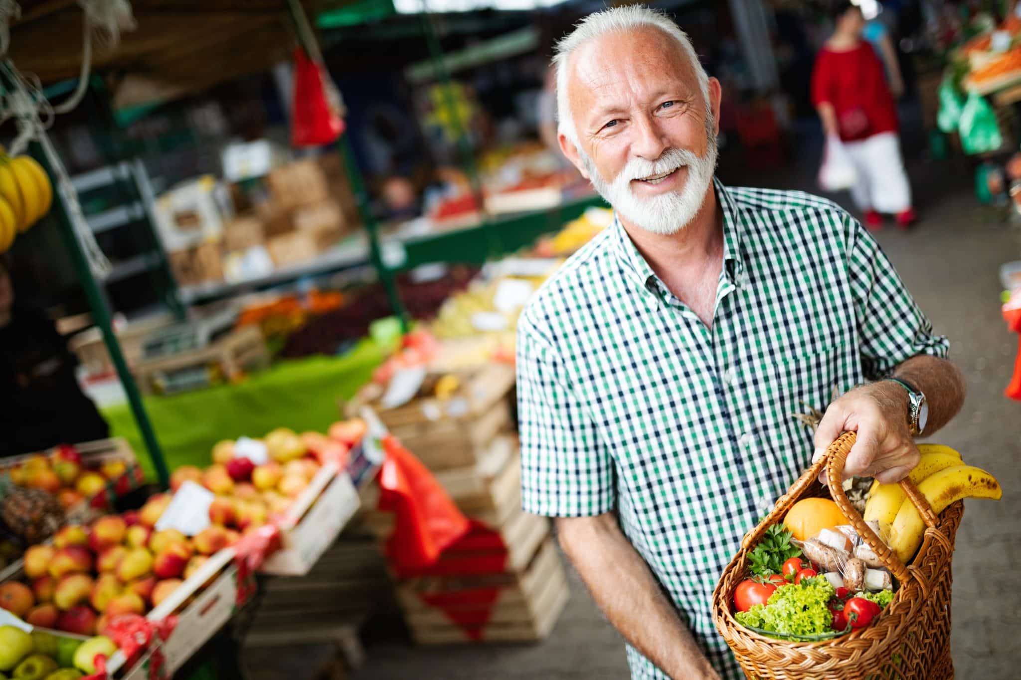 senior man smiling and holding basket of fresh produce while shopping at a market