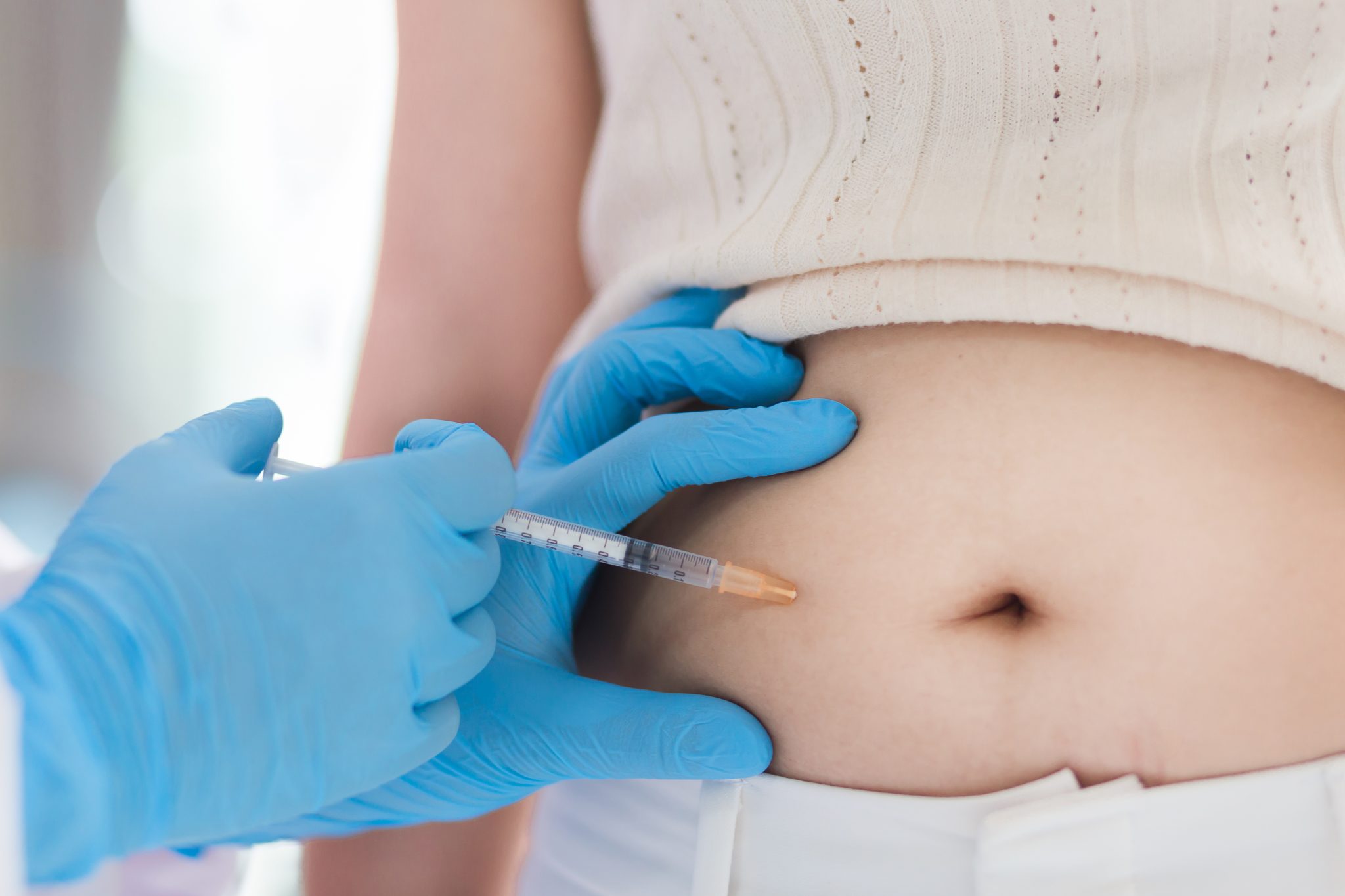 A close-up of a person receiving an injection in their abdomen by a healthcare professional wearing blue gloves.