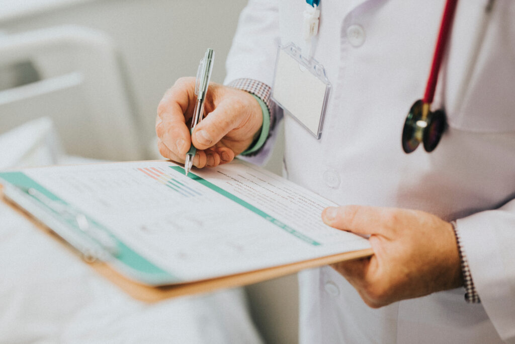 A doctor in a white coat writes on a clipboard, holding a pen and reviewing a medical chart. The doctor’s stethoscope and ID badge are visible, emphasizing a clinical setting focused on patient care.