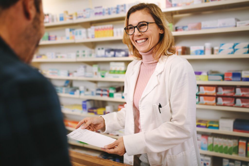 A smiling female pharmacist in a white lab coat hands a prescription to a male customer at a pharmacy counter, with shelves of medicines in the background.