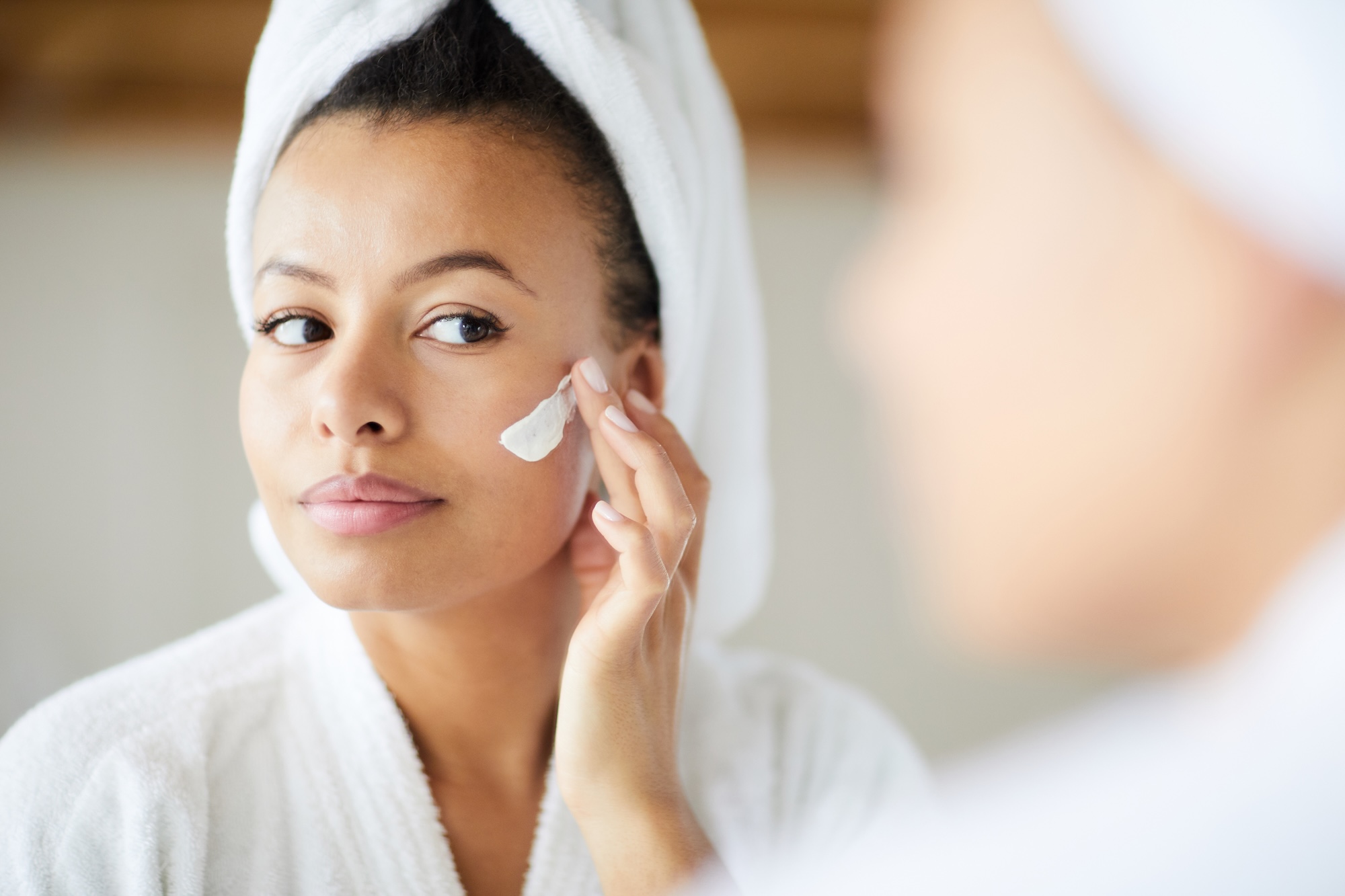 A woman with a towel wrapped around her head is applying skincare cream to her face while looking into a mirror.