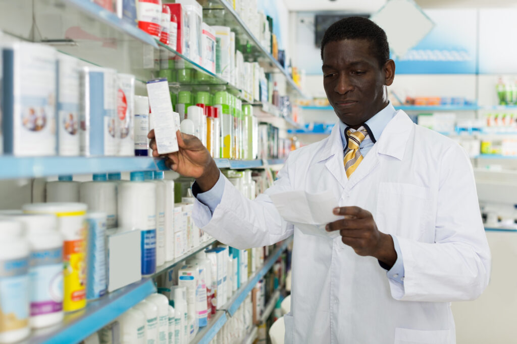 A pharmacist in a white lab coat reads a prescription while selecting a product from a shelf in a pharmacy. The shelves are stocked with various medicines and healthcare items.