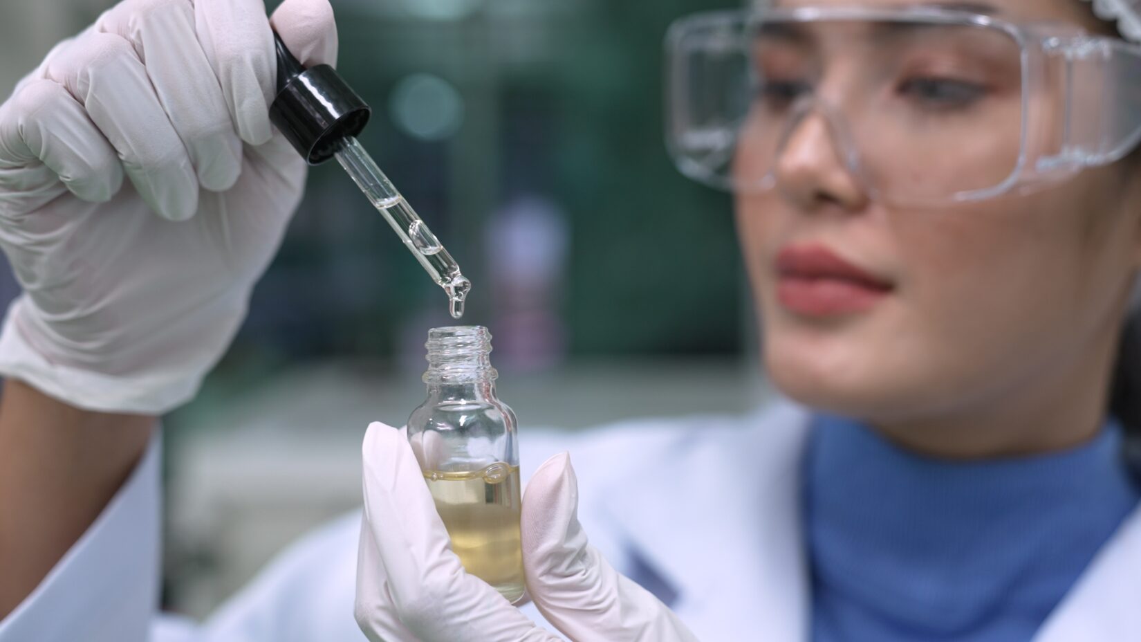 A scientist wearing safety goggles and gloves uses a dropper to carefully add liquid into a small glass vial. The background is blurred, focusing attention on the precision of the experiment.