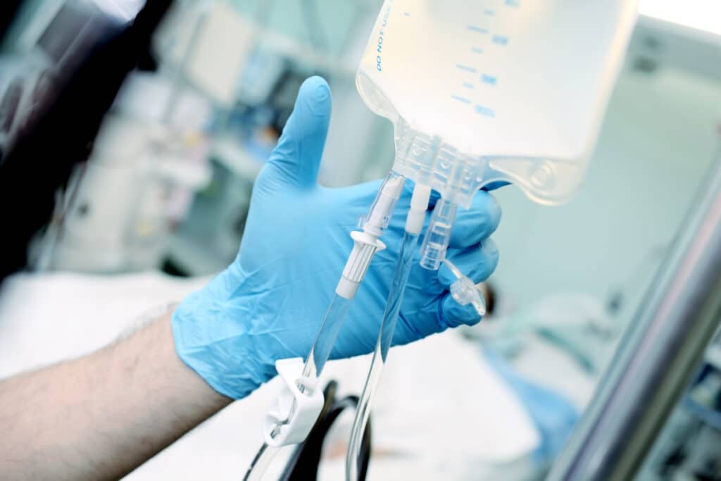 A gloved hand adjusting an IV line connected to a fluid bag in a clinical setting.