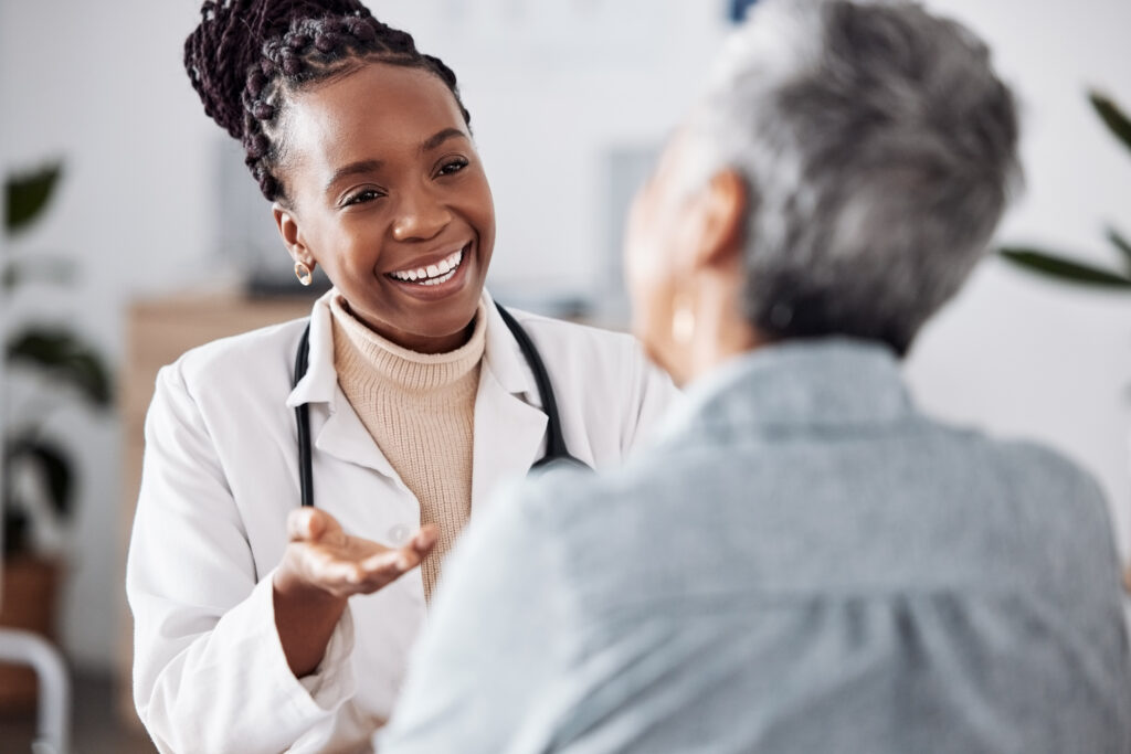 A smiling doctor wearing a white coat and stethoscope engages warmly with a patient during a consultation in a bright office setting.