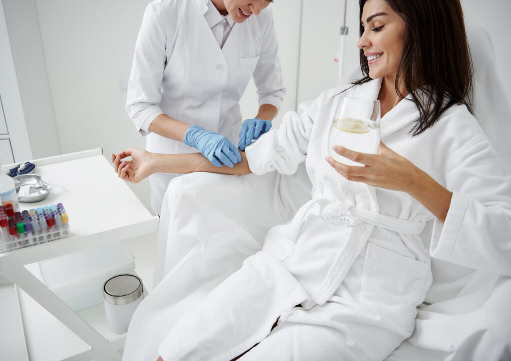 A smiling woman in a white robe relaxes with a glass of water while a healthcare professional in gloves prepares her arm for an IV treatment in a clinical setting.