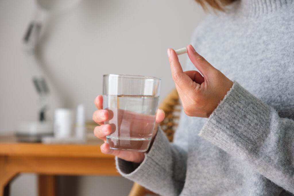 A person in a cozy gray sweater holding a glass of water in one hand and a white capsule pill in the other, preparing to take the medication.