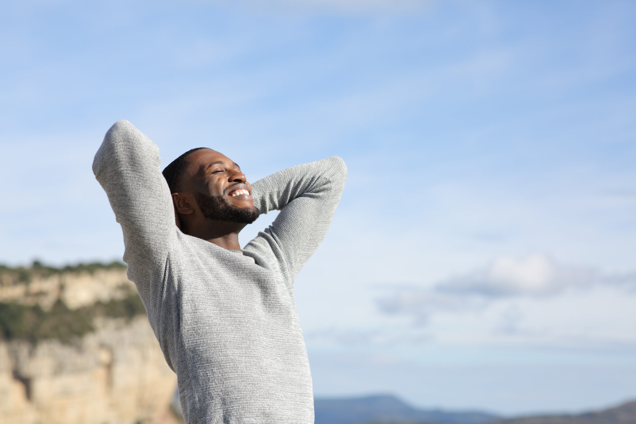 A man in a gray sweater smiles with his eyes closed, stretching his arms behind his head while enjoying the fresh air outdoors with a scenic mountain landscape in the background.