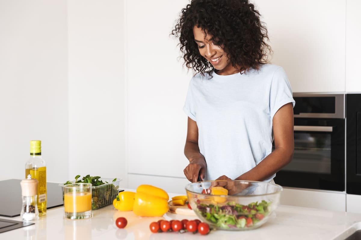 A smiling woman with curly hair prepares a fresh salad in a modern kitchen.