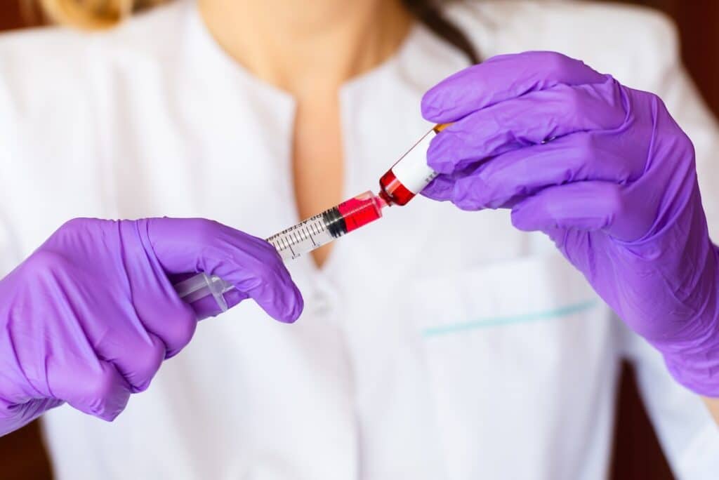 A healthcare professional wearing purple gloves fills a syringe with a red liquid, preparing a B12 injection.