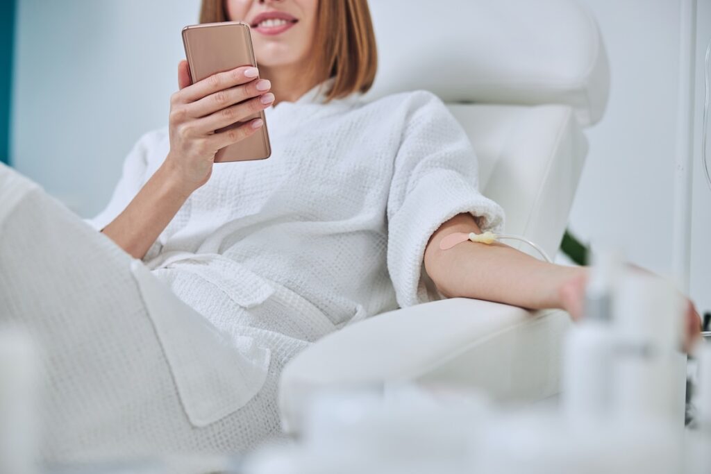 A woman in a white bathrobe is reclining in a chair while receiving an IV drip in her arm. She is holding a smartphone and smiling. The setting is a wellness clinic.